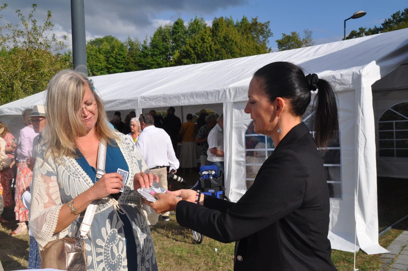 Karine remet une carte à jouer à une spectatrice devant un chapiteau de réception